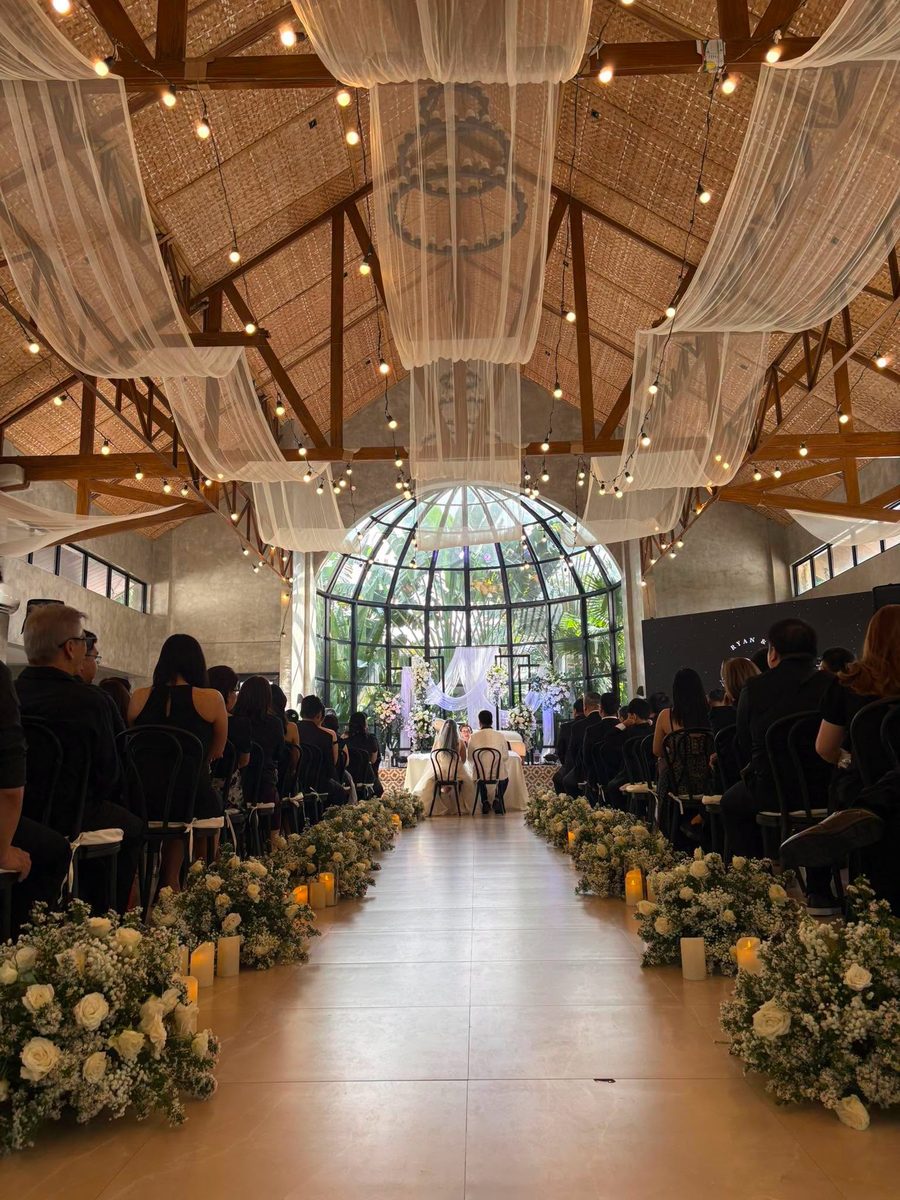 A ceremony aisle lined with white florals and candles under a draped glasshouse roof, coordinated by GO Events.